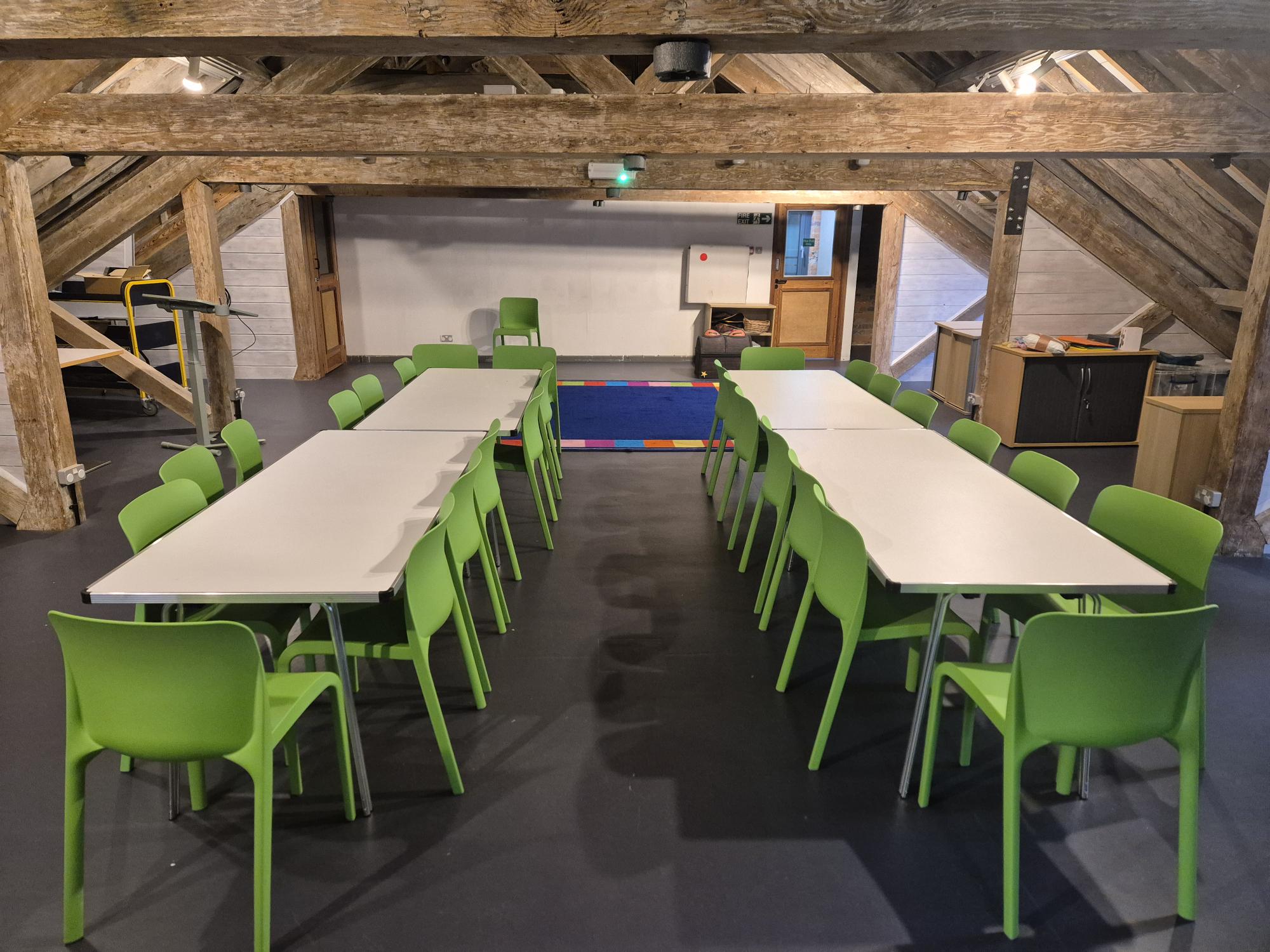A room with tables and chairs lined up, wooden beams and a coloured rug in the background
